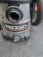 Front close-up of the Ridgid logo on the stainless steel body of the wet/dry vacuum. Hose connection port visible, no suction cup attached.
