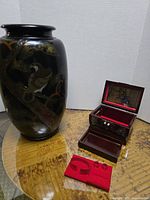 Black oriental vase with bird and floral decoration alongside a small wooden box with mother of pearl inlay and open lid showing red velvet interior and mirror.