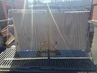 Front view of one clear plexiglass counter shield resting on a wooden board in a vehicle bed under sunlight.