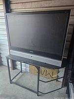 Photo of Samsung large screen TV on metal table stand, shows front view of screen and overall condition.