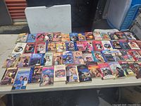 Image showing about 50 vintage romance paperback books laid out on a table, with colorful illustrated covers mainly depicting romantic couples.