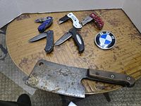 Seven folding knives and one large butcher cleaver knife with wooden handle on a table, plus a round BMW emblem badge.