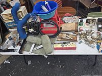 Wide view of table showing blue flexible heater hose, black rubber plug wires, red work stool, seat belts, metal tools and automotive parts spread across the table.