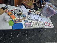 Wide shot of table showing assortment of office supplies, planners, mugs, and miscellaneous items spread out on white folding table outdoors.