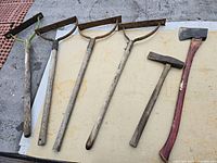 Full view of the lot showing the red-handled ax, hammer ax, hammer, and three grass swing blades laid on a flat surface