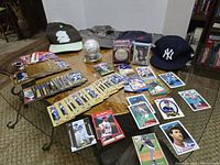 Wide view of baseball memorabilia lot on glass table showing cards, autographed baseballs, and two baseball caps