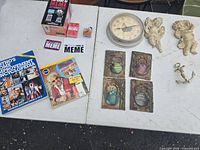 Overview photo of all lot items on table including books, game, clock, tiles, and wall ornaments.