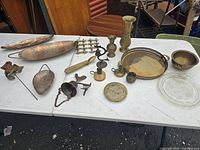 Overview of all brass and copper items on white table including tall vase, trays, bell ringer, candle holders, and decorative pieces