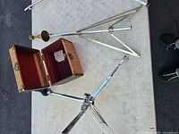 Photo showing vintage camera storage box open with red felt inside, and chrome metal horn stand with tripod base lying next to it