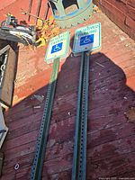 Four reserved handicap parking signs mounted on green perforated metal poles laid out on a wooden surface, viewed from above. Two signs have 'RESERVED PARKING' text with wheelchair symbol in blue and white, two signs have similar but varying design.
