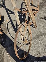 Close-up image showing the rusted metal wheel with thin spokes connected to the rusted metal plow body and weathered wooden handles.