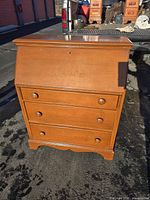 Front view of vintage maple wood secretary desk with three drawers and a closed fold-down writing surface. Shows overall condition with surface scratches and wear.