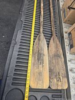 Two vintage wood boat oars laid parallel on a black surface with a measuring tape showing length around 56 inches each. Blades display wear and some weathering.