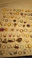 Photo showing approximately 70 assorted rings arranged in rows, featuring various colors and styles including bands, decorative stones, and shapes.