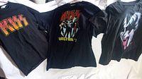Photo of three black KISS T-shirts laid flat showing front designs: classic KISS logo, World Tour '77 with band, Gene Simmons face makeup image.