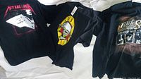 Three black rock band T-shirts laid out flat: Metallica left, Guns N' Roses center, Bob Seger right.