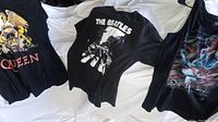 Three rock band T-shirts laid out flat showing front designs: Queen with colorful crest, The Beatles with Abbey Road album cover, and Jimi Hendrix colorful tank top