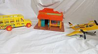 Three vintage toys: yellow Fisher-Price school bus, Fisher-Price Western Town building, and Tonka Radio Commander airplane with propeller, displayed on white background.
