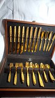 Full set displayed in wood box showing knives, forks, spoons, and butter knives with intricate gold embossing on handles