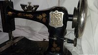 Side view of black fiddlebase sewing machine with gold floral accents, showing hand wheel and brand insignia plate