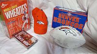 Photo showing the Wheaties cereal box in plastic protector, signed Broncos cap, signed football, unopened John Elway video cassette, and Broncos trivia box game