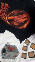 Dungeons & Dragons black T-shirt with orange dragon graphic and text, castle-shaped cookie jar, and Magic The Gathering cards displayed on white surface