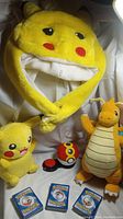 Photo showing yellow Pikachu hat, plush Dragonite, plush Pikachu, Pokeball toy, and three stacks of Pokemon cards
