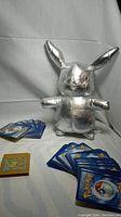 Silver metallic Pikachu plush, multiple stacks of Pokémon trading cards, and yellow Pokémon Gold Game Boy cartridge arranged on a white backdrop.