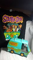 Photo showing Scooby-Doo black T-shirt with colorful Mystery Machine and characters print behind the toy van.