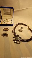 Wide angle showing the sterling silver flower pendant necklace on brown leather cord, moonstone pendant, elephant pin, leather flower pin, and the Colors of Life vintage ring set box with interchangeable stones.