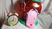 Photo showing the Strawberry Shortcake alarm clock and strawberry-shaped playhouse side by side on a white cloth
