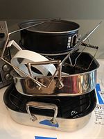 Stack of two All-Clad stainless steel roasting pans with attached handles and three black non-stick cake pans on top. Visible signs of light use.