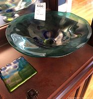 Overhead view of large art glass pedestal bowl showing glossy surface and wavy rim, with small square decorative tile nearby reflecting similar color tones.