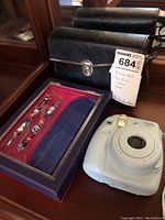 Photo showing black leather case for binoculars, boxed decorative pen set with three pens, and Fuji Instax Mini 8 camera in light blue.