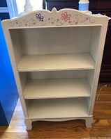 Front view of the white painted bookcase showing two shelves and floral and butterfly painted design on the top edge.
