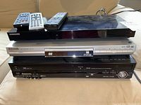 Three stacked players: Samsung 3D Blu-ray player on top, Panasonic DVD player in middle, Panasonic VHS player at bottom with cables visible.