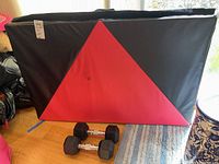 Gymnastics mat folded upright with visible triangular red and black design and two dumbbells in front.