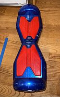 Top view of blue and red hoverboard placed on wood floor, showing red footpads and blue plastic body with visible crack near end.