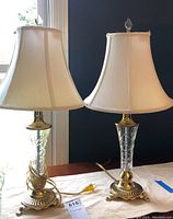 Pair of brass and glass table lamps with cream fabric bell-shaped shades on a wooden surface near window, one lamp shade showing some wrinkles or imperfections.