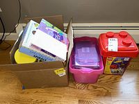 Box filled with various craft and design books and notebooks beside two plastic containers on a wooden floor below a window.