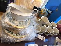 Stack of plates and gravy boat wrapped in plastic showing gold rims and holly leaf patterns.