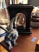View of black mantel clock with glass front showing mechanical movement and Roman numeral dial, alongside resin winged lion figurine with wings extended and roaring expression.