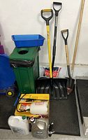 Photo of outdoor supplies including shovels, waste basket, ice melt, Round-up, and fire logs arranged on floor and near steps.