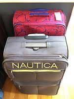 Two rolling soft-side suitcases stacked, the top one is red with an abstract pattern and the bottom one is a brown Nautica suitcase with yellow zippers and logo.