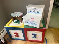 Wooden toy chest with blue and red framing and numbered 2 and 3 compartments, with three stools on top: two round and one step stool painted with sailboat scenes.