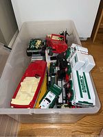 Plastic container with various toy trucks including Hess and Tonka, and a red plastic Tonka speed boat visible on top, inside a plastic bin.