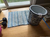 Umbrella stand sitting partially on small blue and white woven rug on wooden floor next to window.
