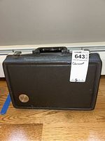 Closed black textured clarinet case with handle and metal clasps, sitting on wooden floor.
