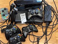 Xbox 360 console with five black wireless controllers, wired controller, clear wired controller, and power adapter on wood floor.
