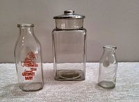 Front view of the three glass items: one large store display jar with aluminum lid, one vintage Syracuse Guernsey Dairy milk bottle, and one smaller Mojonnier Dairy bottle.
